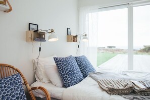 A bedroom with a bed, two wall-mounted lamps, a window with a view, and a wooden shelf with books. - Johanna's Hotel (Bua)