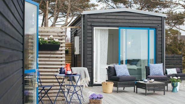 A wooden deck with a small outdoor seating area, a glass door leading to a room, and a wooden structure with a blue trim. - Johanna's Hotel (Bua)