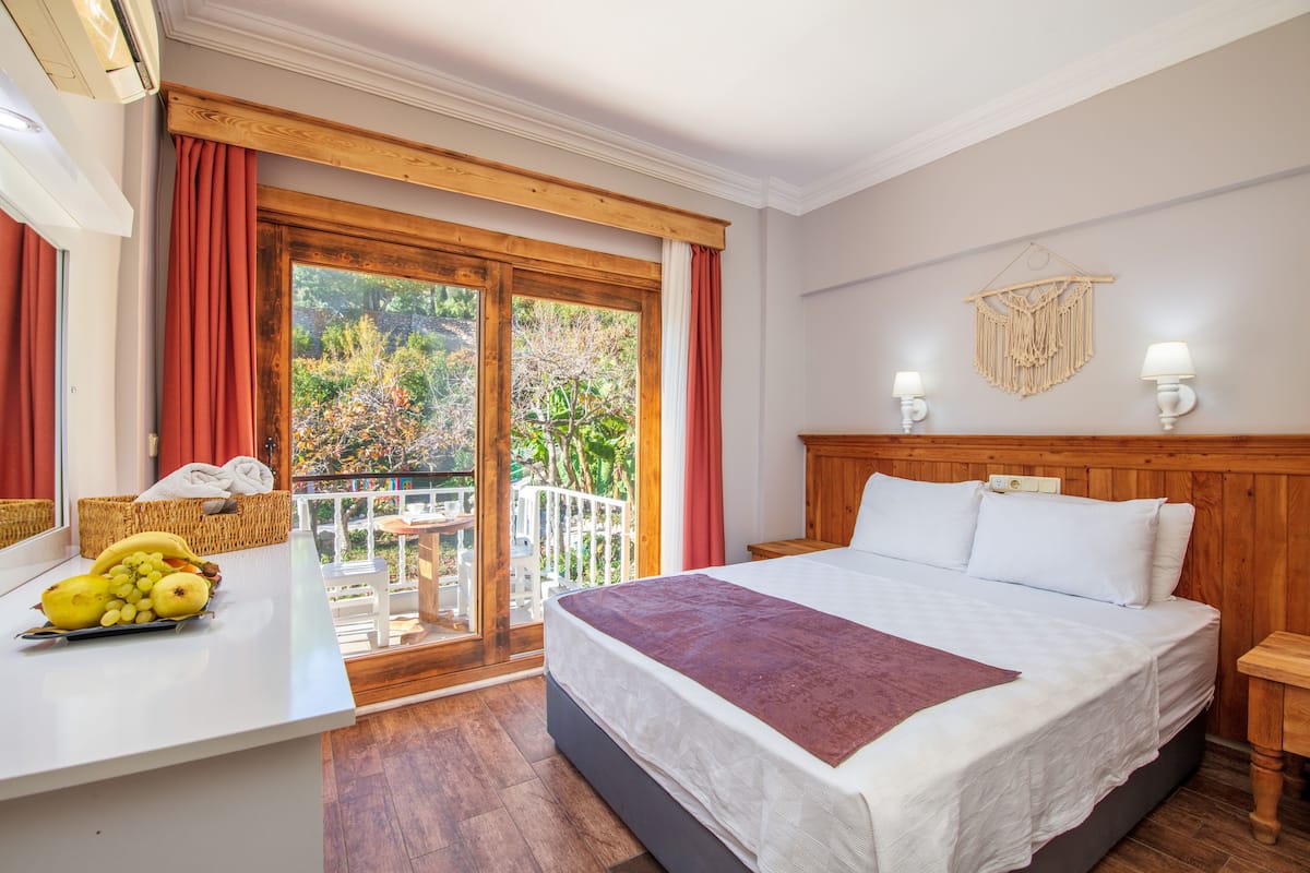 Un dormitorio con cama, una mesita de noche con frutero, ventana con cortinas y balcón con vistas a árboles.