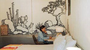 A person sitting at a table in a room with a large wall mural, a bed with white bedding, and a lamp. - Outsite Todos Santos - Work, Connect, Explore (Todos Santos)