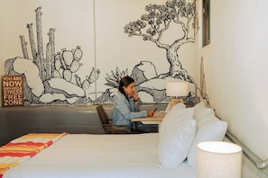 Une personne est assise à une table dans une pièce ornée d’une grande fresque murale, avec un lit recouvert de linge de lit blanc et une lampe.