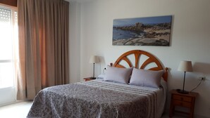 Un dormitorio con una cama, dos mesitas de noche, una mesilla auxiliar, una ventana con cortinas y un cuadro colgado en la pared.