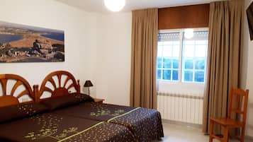 Un dormitorio con cama, una silla, un ventanal con cortinas, un radiador y un cuadro en la pared.