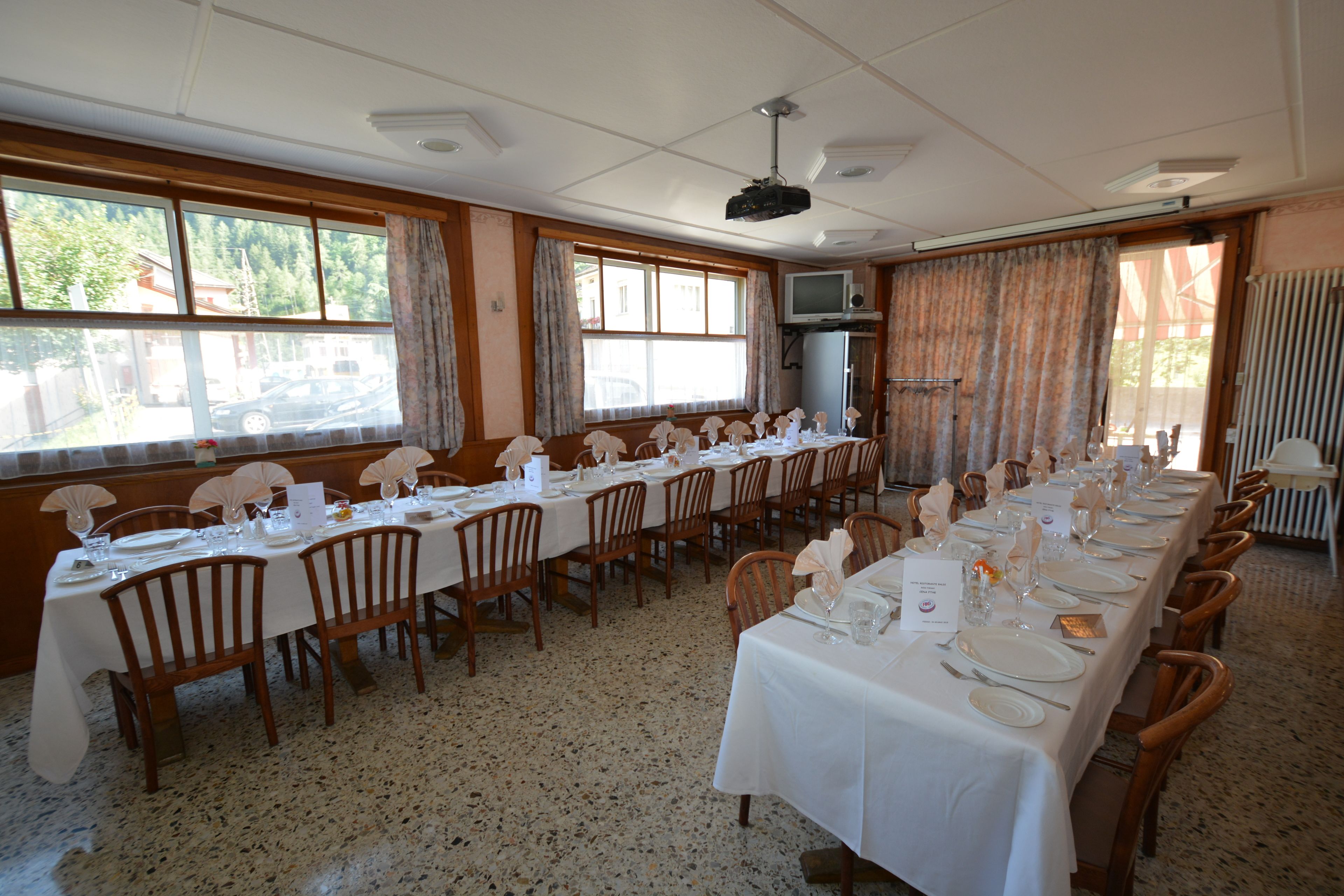 Een lange eettafel gedekt voor een formeel evenement, met stoelen en tafels in een ruime kamer met grote ramen en een plafondprojector.