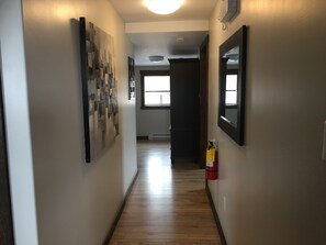 A hallway with wooden flooring, a fire extinguisher mounted on the wall, and artwork on the walls. - Admirals Bed and Breakfast (Twillingate)