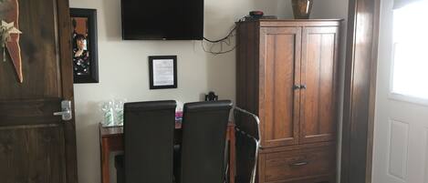 Comedor con mesa de madera, sillas negras, televisor montado en la pared y un armario con un florero encima.