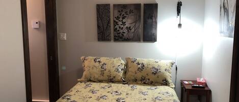 Een slaapkamer met een bloemenbedekte lakens, een nachtkastje met een lamp, en twee aan de muur bevestigde kunstwerken.