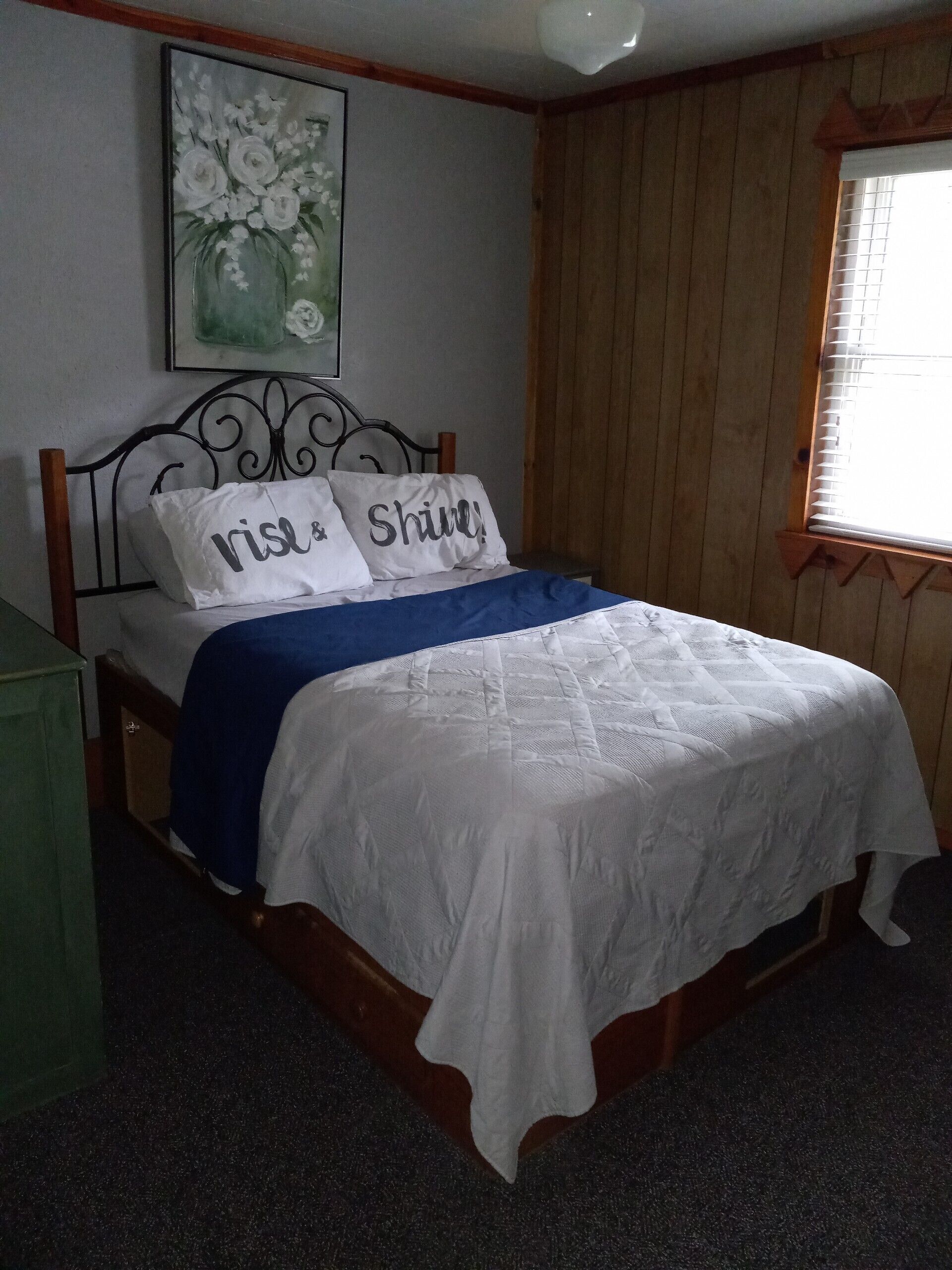A neatly made bed with a white comforter and two pillows, one with "ris" and the other with "shine!" text.