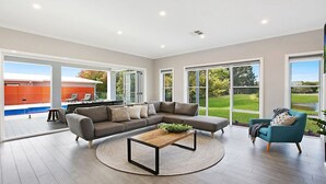 A spacious living room with a large sectional sofa, a coffee table, and a view of a swimming pool and outdoor area through large windows. - CHATEAU CALIZA - The Vintage Holiday Houses (POKOLBIN)