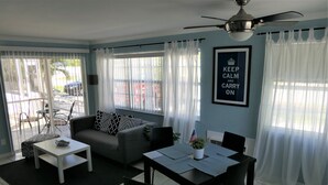 A living room with a sofa, coffee table, dining table, and chairs. There is a ceiling fan, a window with curtains, and a framed sign on the wall. - Sun, beach, and great savings: huge discounts on holiday destinations! (Cape Coral)