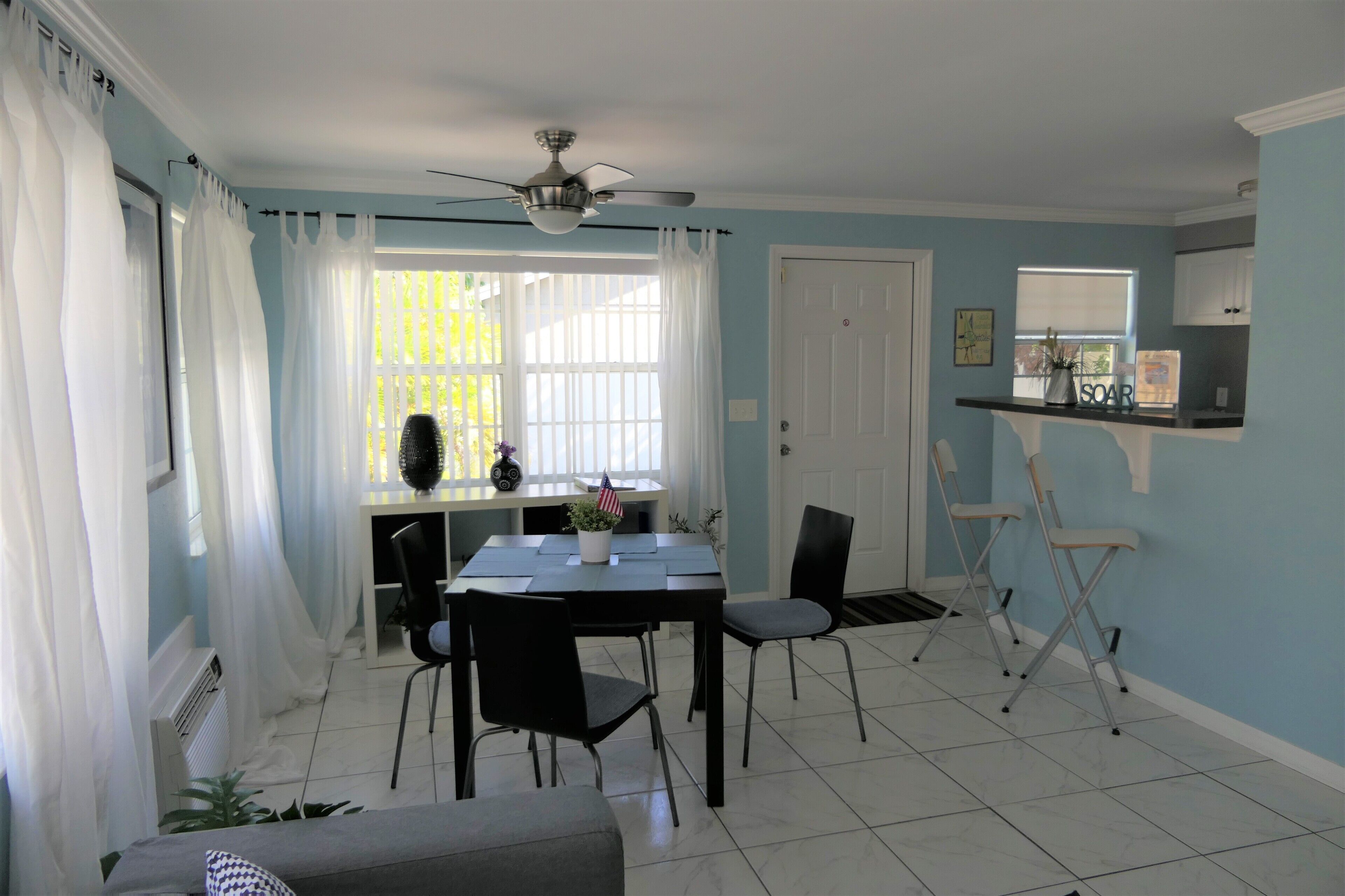 Uma sala de jantar com uma mesa, cadeiras, um ventilador de teto e uma janela com cortinas.