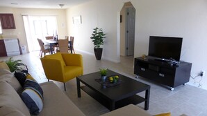 A living room with a sofa, coffee table, TV stand, and a dining area in the background.