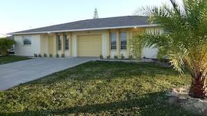 A single-story yellow house with a garage, a driveway, and a small palm tree in the front yard.
