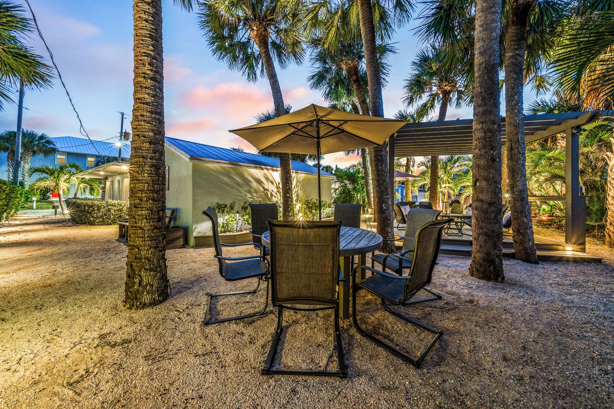 An outdoor seating area with a table and chairs, surrounded by palm trees and a covered structure.