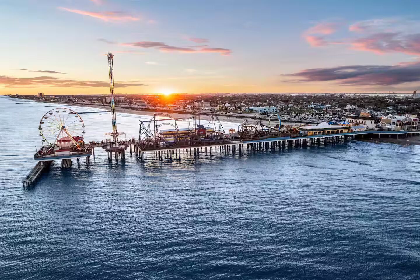 A pier extending into the water with a Ferris wheel and amusement rides, overlooking a coastal cityscape at sunset.