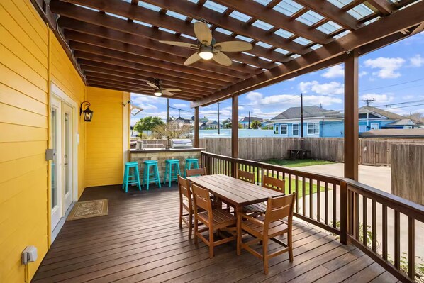 A covered patio with a wooden table and chairs, a ceiling fan, and a view of a residential street.