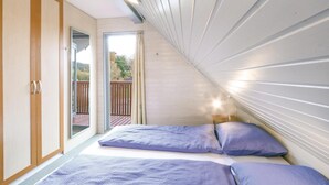 A bedroom with a sloped ceiling, a bed with blue bedding, a wooden wardrobe, and a view of a deck and trees. - Ferienhaus 41 in Kirchheim (Kirchheim/Hessen)