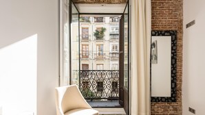 A room with a view of a brick building, a white chair, a wooden table, and a mirror on the wall.