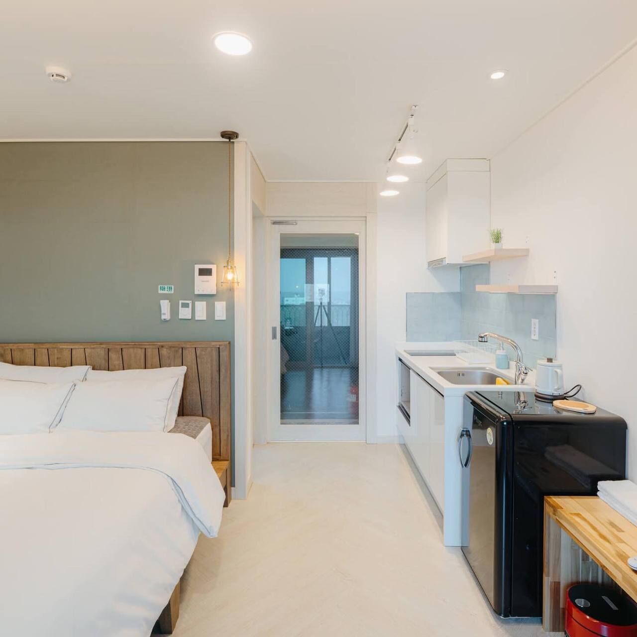 A compact hotel room with a bed, kitchenette, and a view through a glass door.