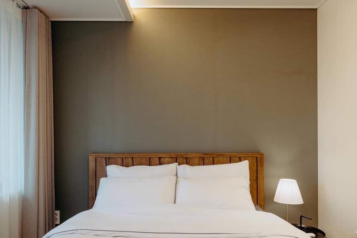 A neatly made bed in a hotel room with a wooden headboard, a bedside lamp, and a view of the window with curtains.