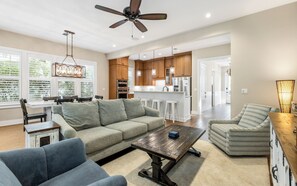 A modern living room with a sofa, coffee table, and chairs, a kitchen with wooden cabinets and stainless steel appliances, and a ceiling fan.