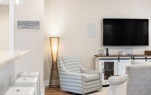A living room with a checkered armchair, a floor lamp, a TV, and a cabinet with books. - Sunseeker - Seagrove Home + Pools, Grill & Bikes! (Santa Rosa Beach)