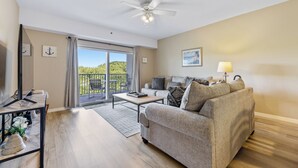 A living room with a sofa, coffee table, and a ceiling fan. There is a sliding glass door leading to a balcony with a view of greenery.