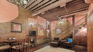 A cozy living room with exposed brick walls, a wooden ceiling, and a staircase. There is a TV on a wooden stand, a round table with chairs, and a patterned rug. - Crossroads Loft:  Steps from Rt. 66 & Carthage Square (Carthage)