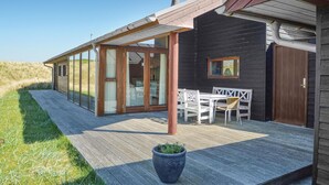 Ein modernes Holzhaus mit einer glasvergrößerten Terrasse, einem Essbereich mit weißer Möbel und einer großen Topfpflanze im Vordergrund.