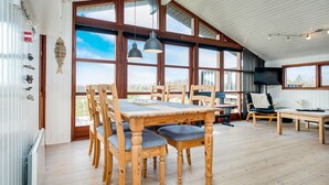 A spacious room with a large wooden dining table, multiple chairs, a television, and large windows offering a view of the outdoors. - Gorgeous home in Løkken with kitchen (Løkken)