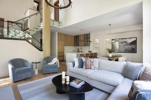 Uma sala de estar moderna com escada, um sofá, poltronas, uma mesa de centro e uma cozinha ao fundo.
