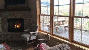 A cozy living room with a fireplace, a comfortable chair, and a large window overlooking a deck with outdoor furniture. - Yellowstone Archview Guesthouse- Stunning Views For Your Ultimate Family Getaway (Gardiner)