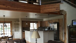 A spacious living room with a brown leather sofa, a dining table with chairs, and a kitchen area with wooden cabinets and a stainless steel refrigerator. - Yellowstone Archview Guesthouse- Stunning Views For Your Ultimate Family Getaway (Gardiner)