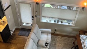 A living room with a white sofa, a dining table with chairs, a window with blinds, and a wall-mounted light fixture. - University Park Garden Guest House (Portland)