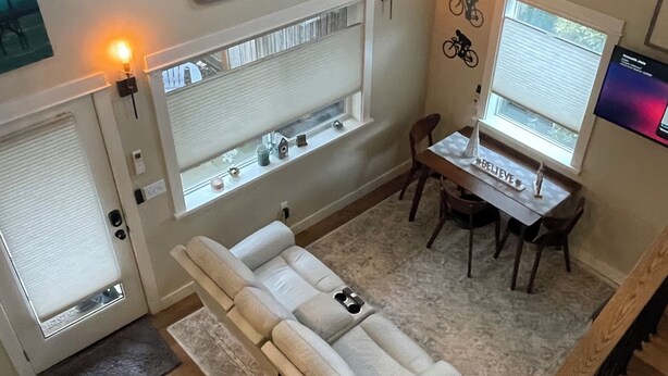 A cozy living area with a sofa, a dining table, and a TV. There are two windows with blinds, a fireplace, and a wall-mounted artwork. - University Park Garden Guest House (Portland)