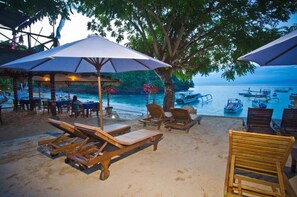 A beachside setting with wooden lounge chairs and umbrellas, a dining area, and a view of the sea with boats. - Lumbung Bali Huts (Lembongan Island)