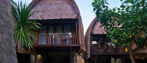 A traditional thatched-roof bungalow with a balcony, surrounded by tropical vegetation.