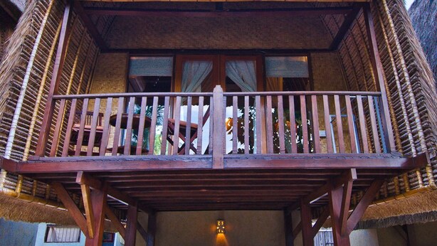 A two-story thatched-roof building with a balcony, wooden furniture, and a small light fixture. - Lumbung Bali Huts (Lembongan Island)