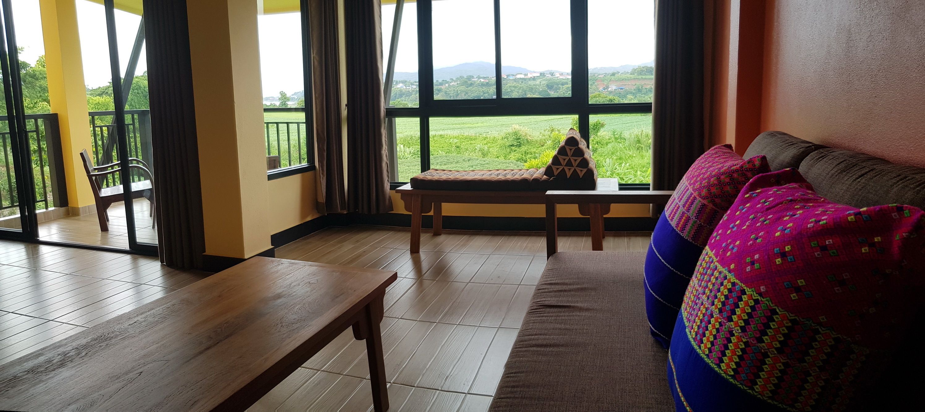 A room with a wooden bench, a sofa with patterned cushions, and a view of a green landscape through large windows.