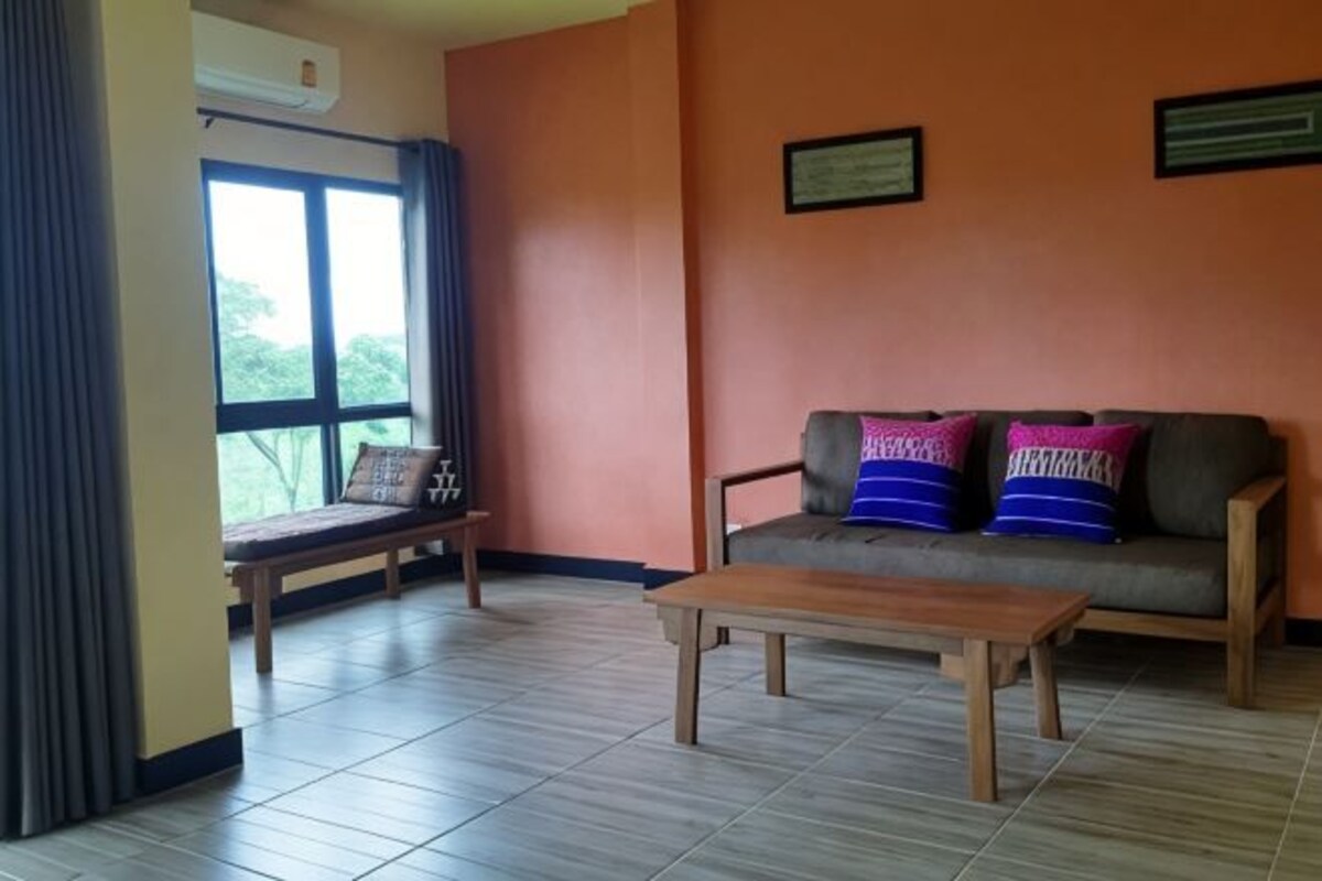 Una habitación con sofá, una mesa de madera, un banco y una ventana con cortinas.