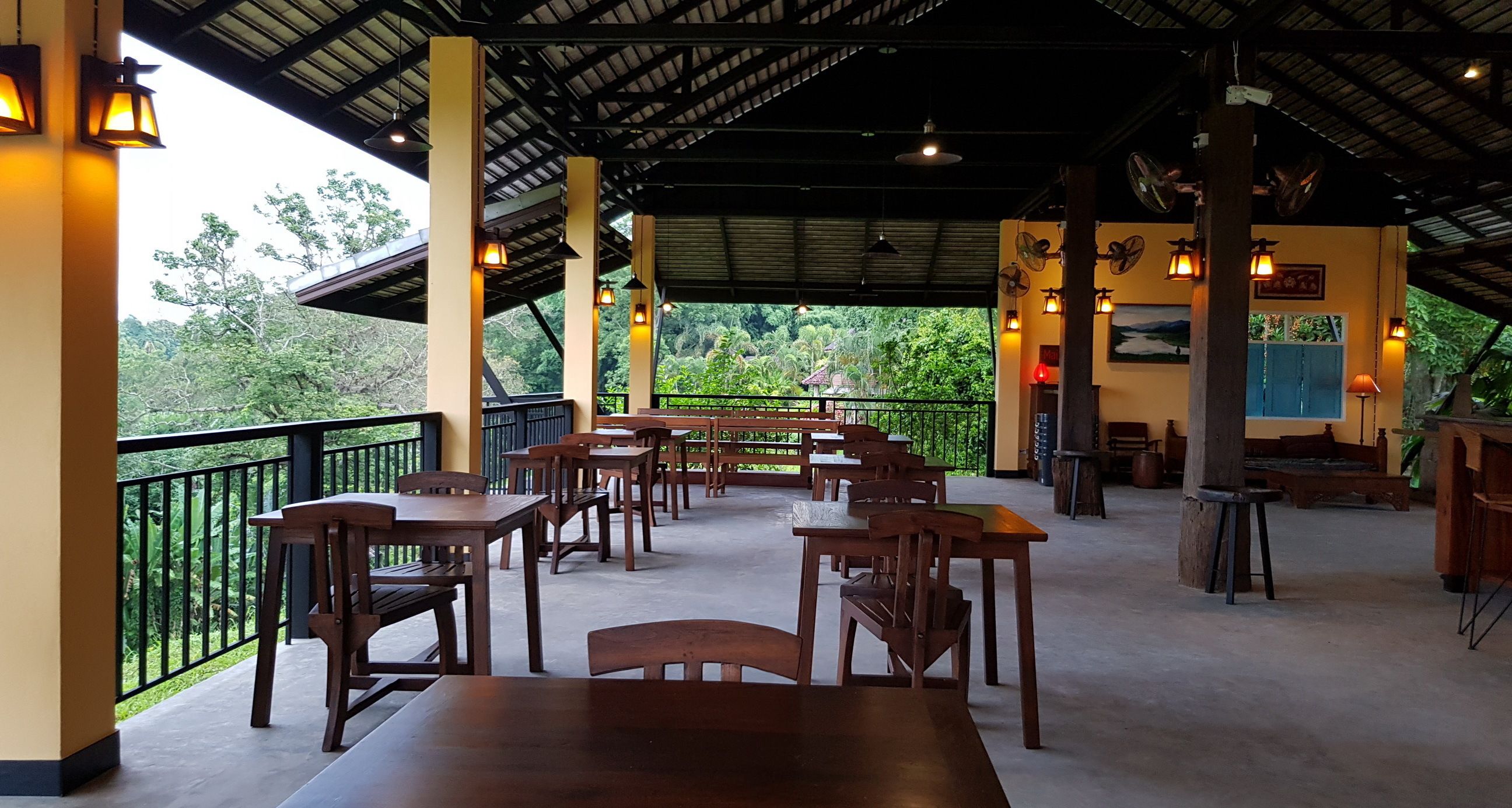 A covered outdoor dining area with wooden tables and chairs, a black metal railing, and a view of lush greenery.