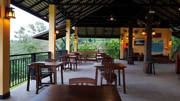 A covered outdoor dining area with wooden tables and chairs, a black metal railing, and a view of lush greenery.