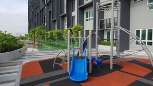 A playground with a blue slide and climbing structures, surrounded by a landscaped area with trees and a building in the background.