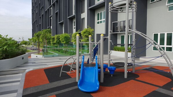 Un parque infantil con un tobogán azul y estructuras para escalar, rodeado de un área ajardinada con árboles y un edificio al fondo.