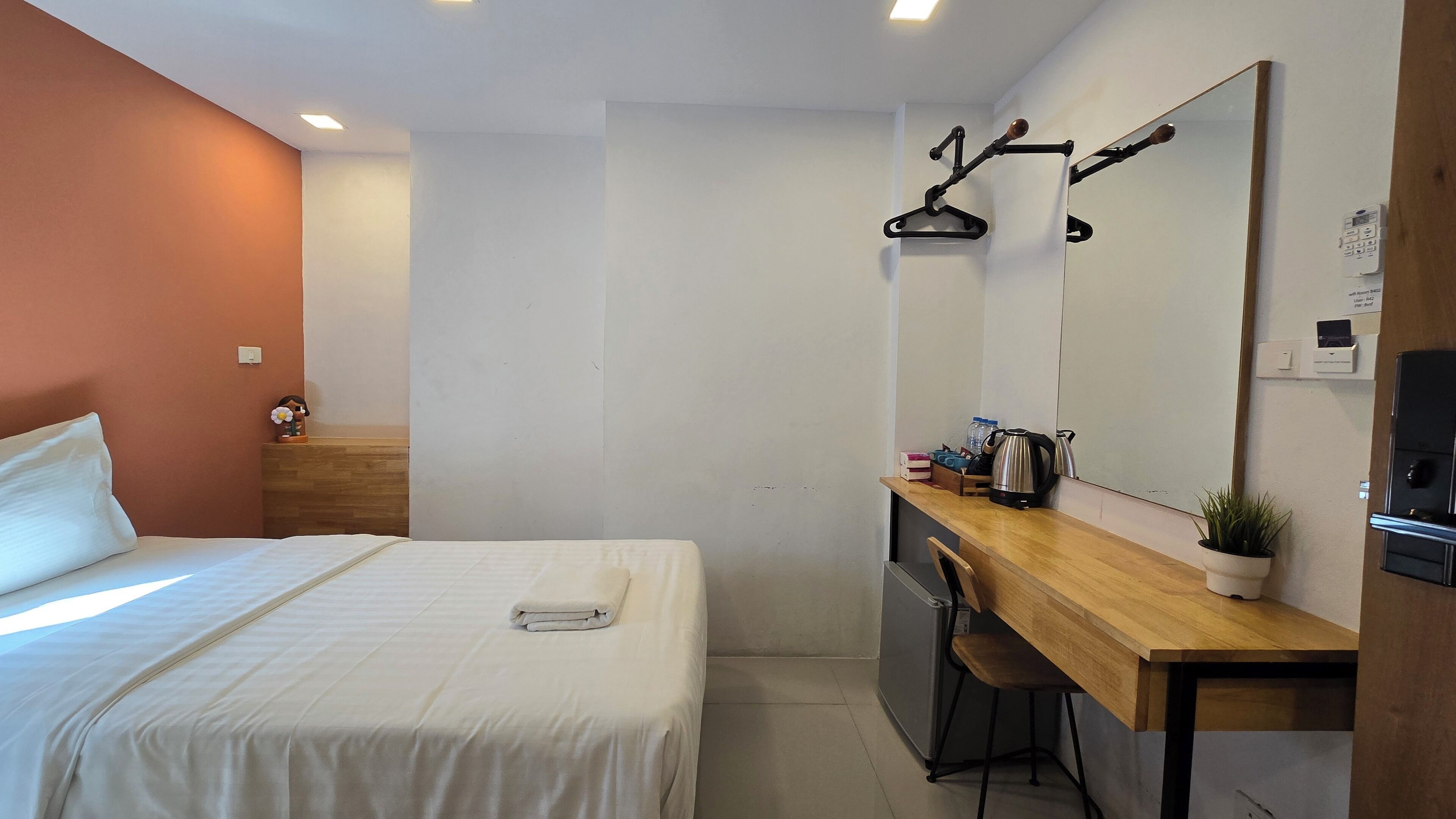 A hotel room with a bed, a wooden desk, a chair, a mirror, a kettle, and a small potted plant.