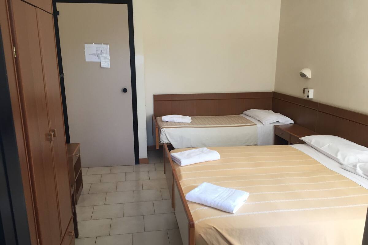 Una habitación de hotel con dos camas, un armario de madera, un escritorio y una lámpara fijada a la pared.