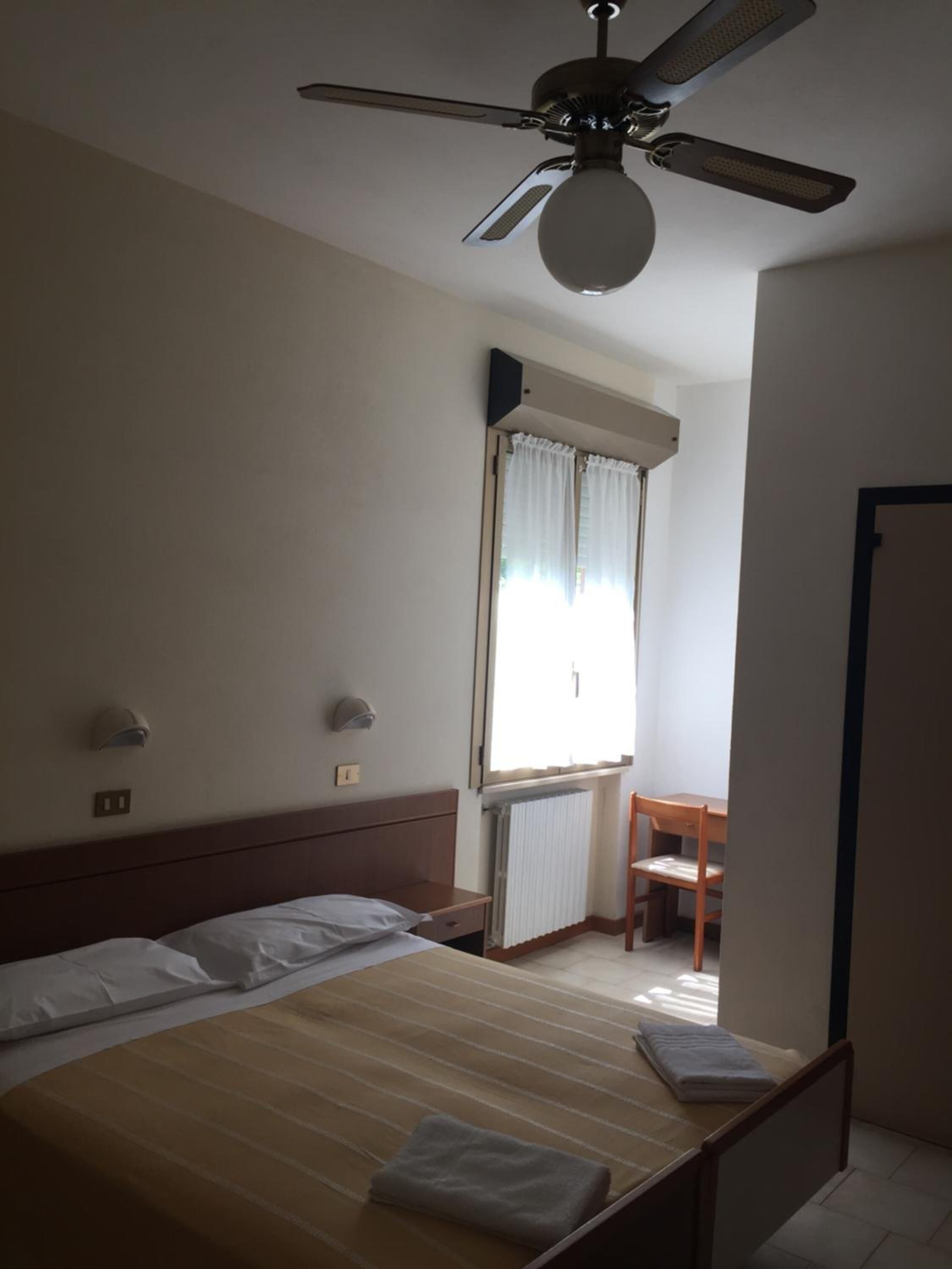 Habitación de hotel con una cama individual, ventilador de techo, ventana con cortinas, radiador y una silla.