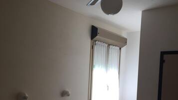 Habitación de hotel con una cama individual, ventilador de techo, ventana con cortinas, radiador y una silla.