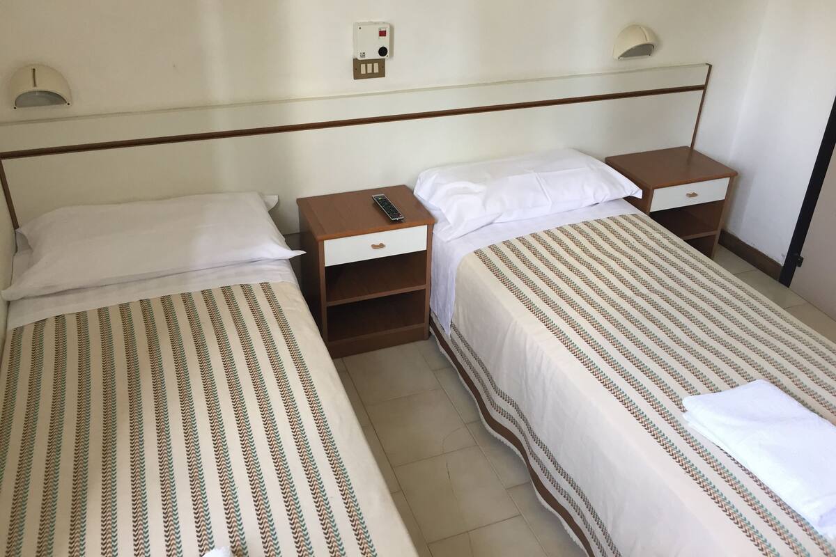 Habitación de hotel con dos camas individuales, cada una con una mesita de noche de madera, un control remoto y un enchufe de pared.