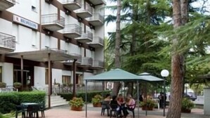 An outdoor seating area with tables and chairs, surrounded by greenery and a building with multiple balconies. - Hotel Belvedere (Castrocaro Terme)